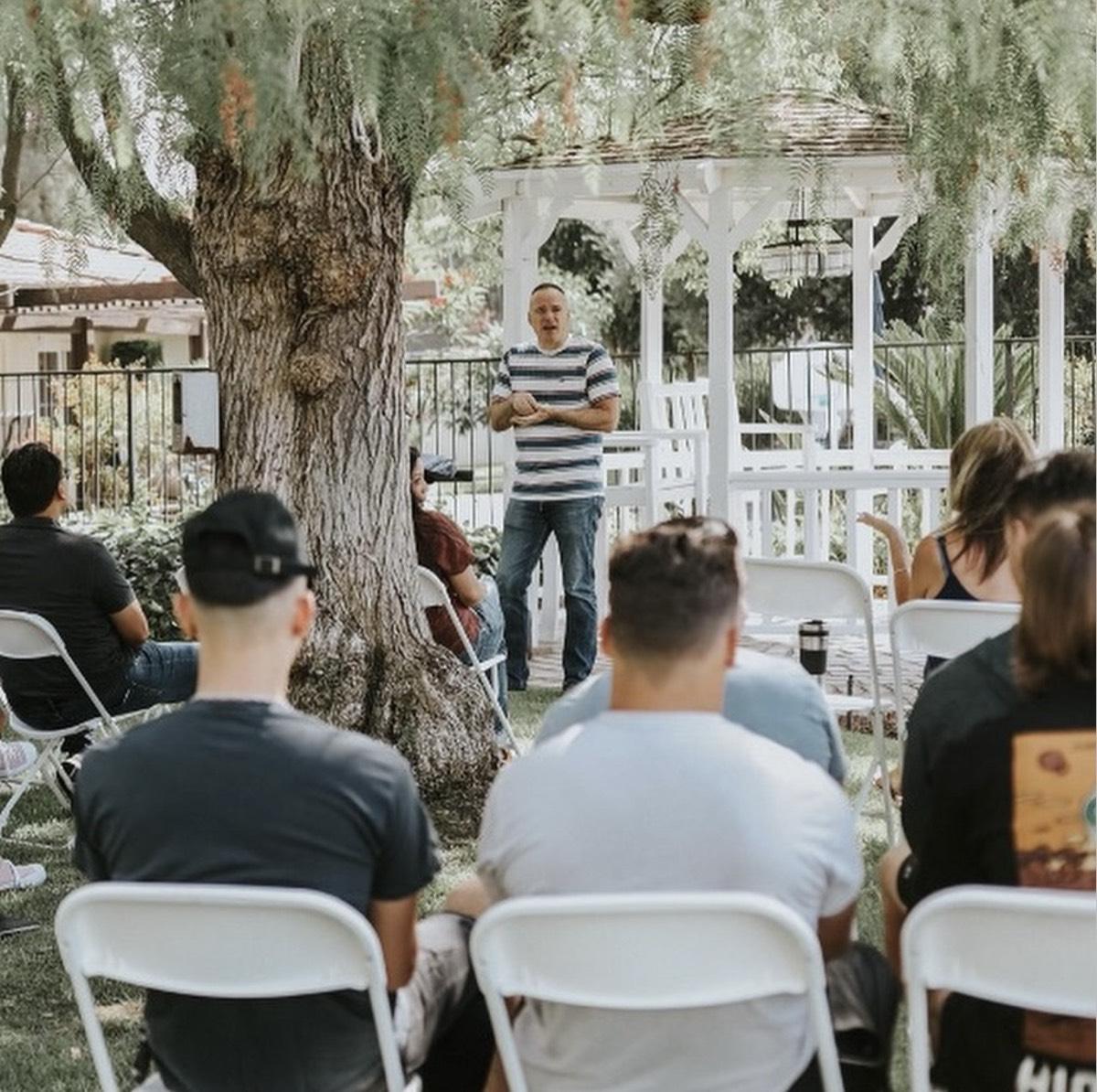 A New Life Church gathering outdoors, listening to a message under a tree